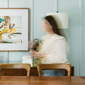 Woman sitting at a wooden table in a room with plants and a framed picture on the wall.